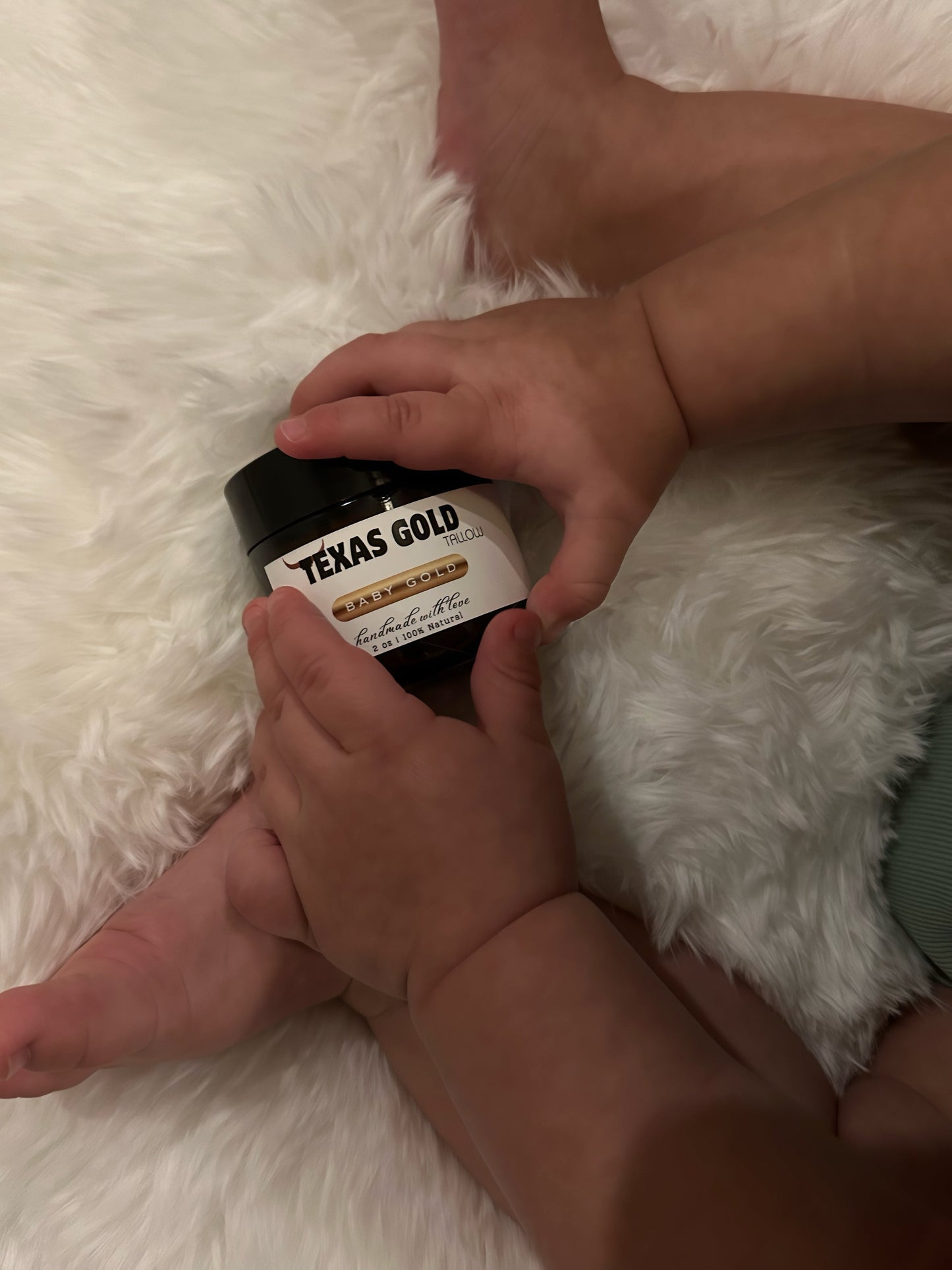 Baby holding Texas Gold Tallow, baby gold balm on a soft blanket, natural grass fed and grass finished beef tallow baby balm for sensitive skin, dry skin, and gentle baby skin care